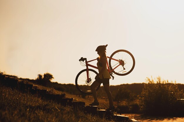 Silhouette einer Frau, die ein Fahrrad gegen den Abendhimmel trägt