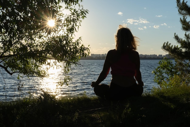 Silhouette einer Frau, die im Sommer in einer Lotusposition am Ufer eines Sees sitzt Meditationssport