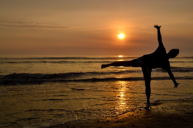 Silhouette einer Frau mit offenen Armen und auf einem Fuß am Ufer eines Strandes bei Sonnenuntergang