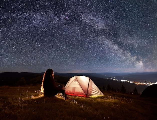 Silhouette einer jungen Dame, die am Feuer und am Zelt unter einem schönen Himmel mit vielen Sternen und Milchstraße sitzt und die Landschaft der Berge und der Stadt entfernt genießt.