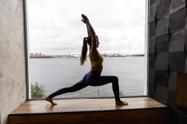 Silhouette einer jungen Frau, die Yoga in Heldenpose macht. Konzept des Quarantäne-Heimtrainings. Werbung