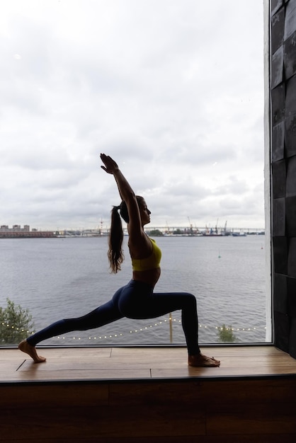 Silhouette einer jungen Frau, die Yoga in Heldenpose macht. Konzept des Quarantäne-Heimtrainings. Werbung