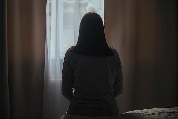 Silhouette einer jungen Frau im Schlafanzug vor dem Hintergrund eines Fensters zu Hause auf einem Bett