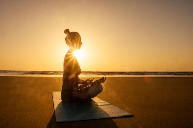 Silhouette einer jungen Frau in einem stilvollen Anzug für einen Yogi-Overall, der Yoga am Strand in Pose-Kopierraum macht.