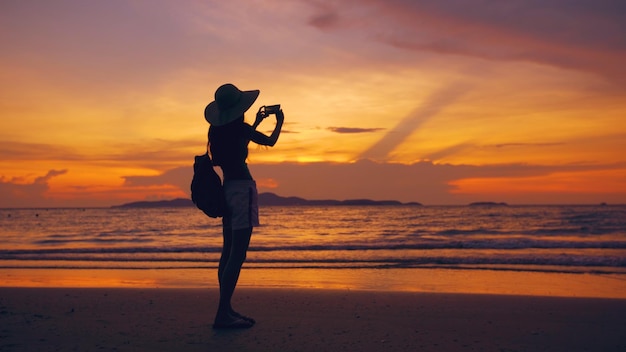 Silhouette einer jungen Touristenfrau mit Hut, die während des Sonnenuntergangs am Meeresstrand ein Foto mit dem Handy macht