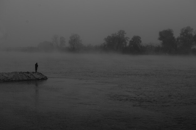 Foto silhouette einer person, die in nebligem wetter in der dämmerung am see auf einem felsen steht