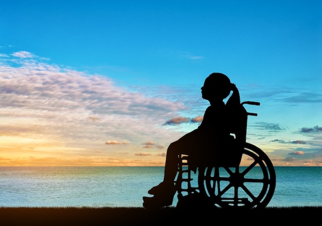 Silhouette eines behinderten Kindermädchens, das in einem Rollstuhl sitzt und einen Meeressonnenuntergang betrachtet. Begriffsbild eines Mädchens mit Behinderung