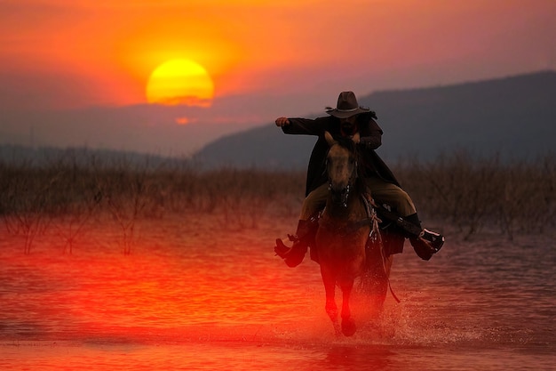 Silhouette eines Cowboys, der auf einem Pferd reitet, das bei Sonnenuntergang hinter einem Berg durch das Wasser watet