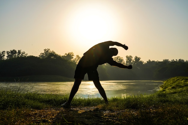 Silhouette eines Jungen, der Übungen im Park in der Nähe von Sonne und Fluss macht - Gesundheitskonzept