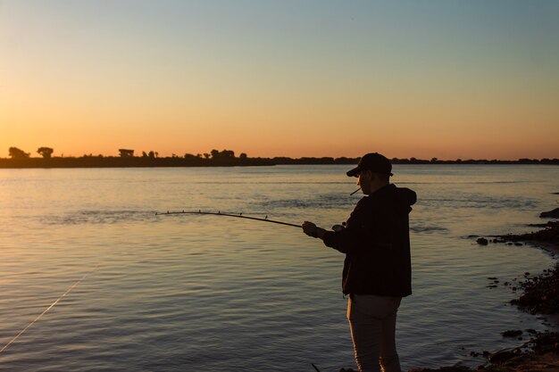 Silhouette eines jungen Fischers, der eine Zigarette raucht und sich am Ufer des Flusses entspannt.