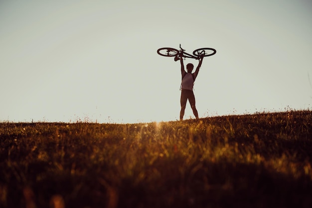 Silhouette eines jungen Mannes, der bei Sonnenuntergang ein Fahrrad auf dem Feld hält, mit einem dramatischen Himmel im Hintergrund Hochwertiges Foto