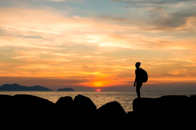 Silhouette eines jungen Mannes mit Rucksack, der auf einem Felsen steht und bei Sonnenuntergang einen schönen Blick auf den Sonnenuntergang über dem Meer hat