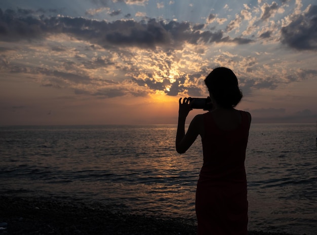Silhouette eines Mädchens fotografiert den Sonnenuntergang am Meer per Smartphone.