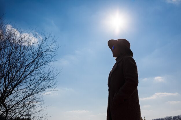 Silhouette eines Mädchens in Hut und Mantel auf dem Hintergrund des Himmels mit Wolken und Sonne.