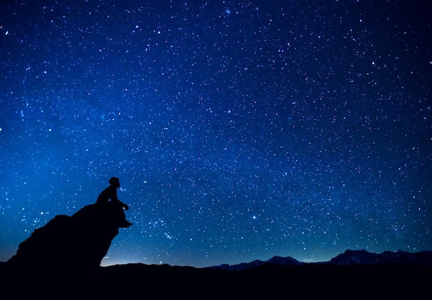 Silhouette eines Mannes, der auf einem Felsen gegen ein Sternbild am blauen Himmel sitzt