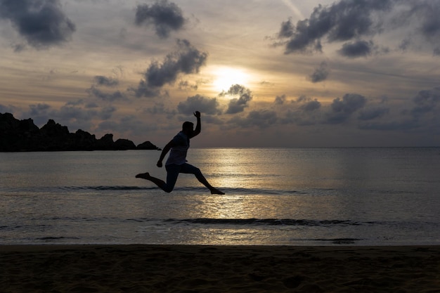 Silhouette eines nicht erkennbaren jungen Mannes, der am Strand springt und glücklich ist, mit dem Sonnenuntergang im Hintergrund