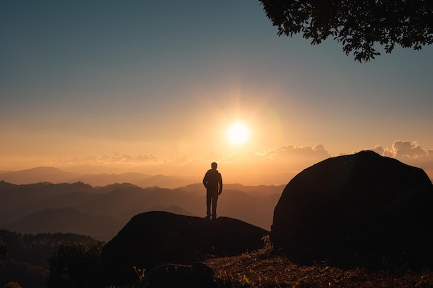 Silhouette eines Wanderers, der auf dem Gipfel eines Berges steht und bei Sonnenuntergang die Sonne über dem Berg scheint