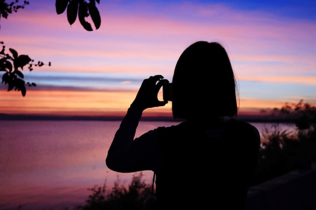 Silhouette Frau fotografiert das Meer mit Smartphone gegen den Himmel bei Sonnenuntergang