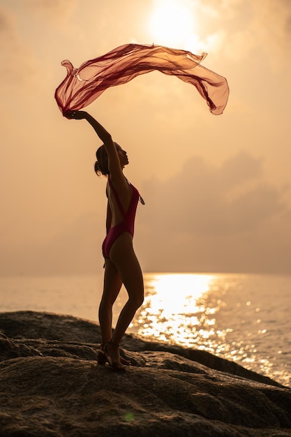 Silhouette Frau mit rosa Badeanzug mit Schal im Wind genießen Sie einen romantischen Sonnenuntergang am steinernen tropischen Strand