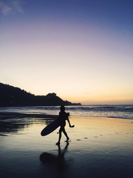 Silhouette Frau mit Surfbrett geht am Strand gegen den Himmel bei Sonnenuntergang