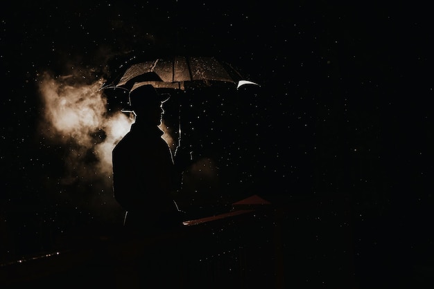 Silhouette im Noir-Stil eines männlichen Mafia-Detektivs in Regenmantel und Hut unter einem Regenschirm