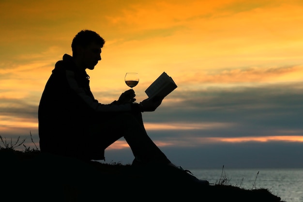 Silhouette Kerl sitzt am Wellenbrecher am Abend in der Nähe von Meer, liest Buch und trinkt Wein