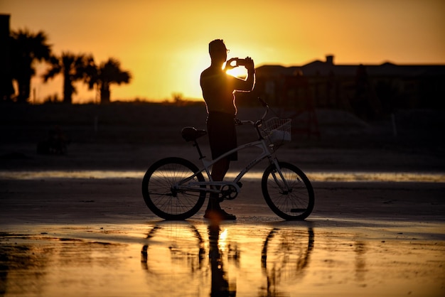 Silhouette-Mann fotografiert mit Mobiltelefon, während er bei Sonnenuntergang am Strand am Fahrrad steht