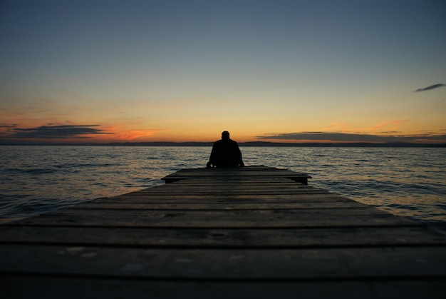 Silhouette-Mann sitzt bei Sonnenuntergang auf dem Steg gegen das Meer