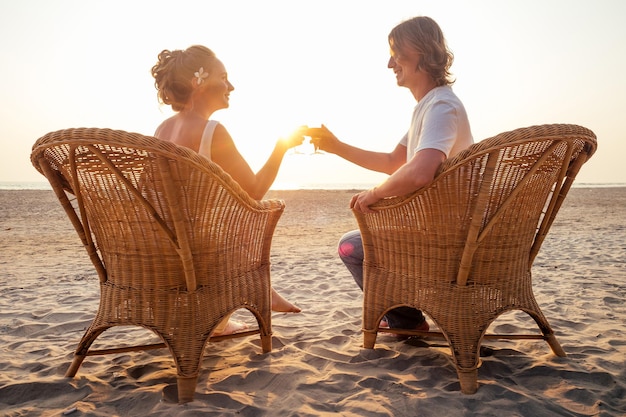 Silhouette verliebtes Paar sitzt auf Korbstühlen und klirrt mit Getränken am Strand mit Blick auf den Sonnenuntergang Valentinstag,