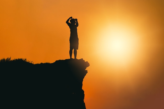 Silhouette von Erfolg und Gelassenheit junger Mann, der bei Sonnenuntergang auf einem Felsen in den Bergen steht.