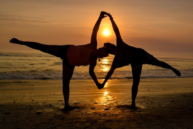 Silhouette von zwei Frauen, die Händchen halten, die bei Sonnenuntergang auf einem Fuß an der Küste stehen