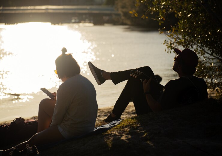 Silhouette von zwei Freunden am Flussufer, die an einem sonnigen Tag Gitarre im Park spielen. Urlaubskonzept