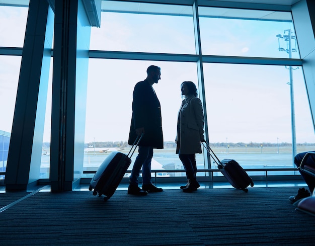 Silhouette von zwei Personen, Frau und Mann mit Koffern, die an den Panoramafenstern mit Blick auf die Landebahnen und Flugzeuge im Abflugterminal des internationalen Flughafens stehen und auf einen Flug warten