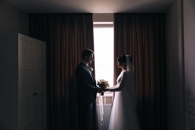 Silhouetten der Braut und des Bräutigams im Innenraum des Hotels in der Nähe des Fensters Hochzeitsporträt von Jungvermählten