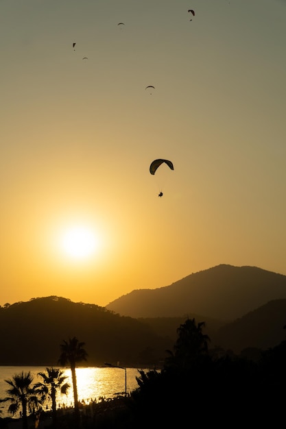 Silhouetten von Gleitschirmen, die bei Sonnenuntergang am Himmel fliegen, wobei das Meer das Sonnenlicht und die Berge im Hintergrund reflektiert