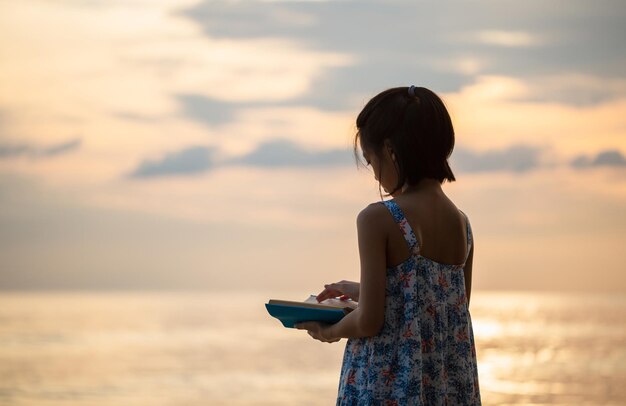 Silhouetten von kleinen süßen Mädchen, die am Strand spielen, Kindermädchen im Kleid, das ein Buch am Strand Sonnenuntergang am Abendhimmel liest