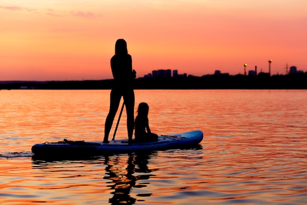 Silhouetten von Mädchen und Kindern, die bei Sonnenuntergang auf Paddle Board paddeln