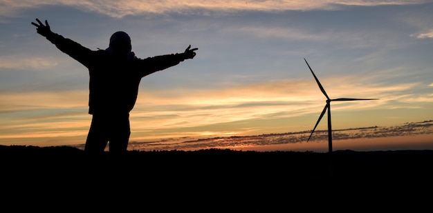 Silhouettieren Sie glücklichen Mann mit den Händen oben und Windkraftanlage in der Sonnenaufgangzeit.