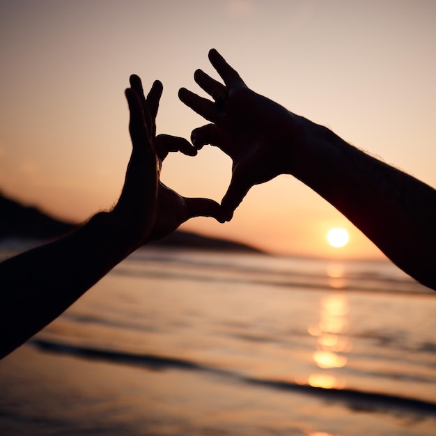 Silhueta da natureza do pôr do sol na praia e pessoa com mãos de coração assinam para amor, liberdade e férias no oceano Ícone de saúde emoji de aventura e apoio de casal com cuidado ou forma tropical de bem-estar natural