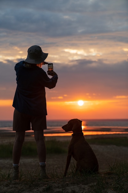 Silhueta de cachorro fêmea e vizsla em pé na praia do mar tirando foto do pôr do sol no celular