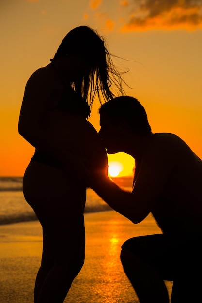 Foto silhueta de homem beijando o abdômen de uma mulher grávida na praia durante o pôr do sol