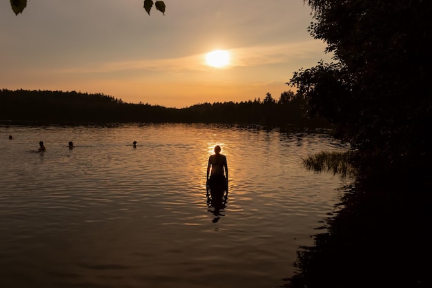 Silhueta de homem em pé e pessoas no lago em reflexo do lago noturno do pôr do sol entre árvores