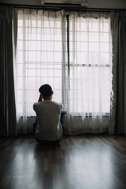 Silhueta de homem sozinho olhando para a janela fechada com cortinas no quarto Homem fica na janela sozinho