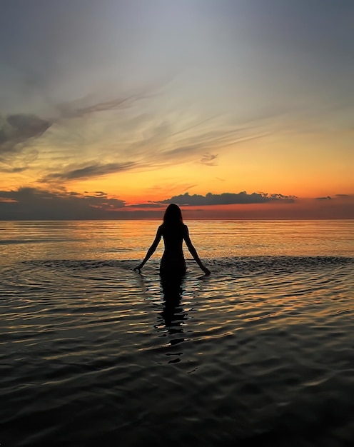 Silhueta de jovem ao pôr do sol laranja na onda de água do mar e céu nublado rosa