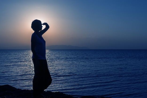 Silhueta de jovem olhando para a distância em pé na praia ao pôr do sol contra o sol