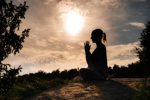 Silhueta de jovem yogini meditando em posição de lótus com os olhos fechados Senhora relaxada está realizando pose de ioga namastê fundo de sol ao ar livre no parque fora da cidade à noite durante o pôr do sol