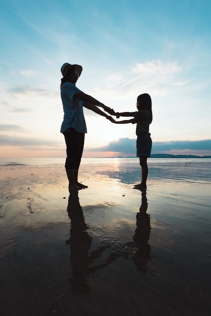 Silhueta de mãe asiática e filha segurando a mão e brincando na praia juntos na hora do sol com lindo mar e céu. Família desfrutar com o conceito de natureza.