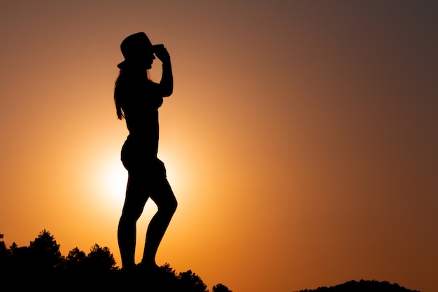 Foto silhueta de mulher de perfil ao pôr do sol de frente para o sol segurando um chapéu na mão copie o espaço