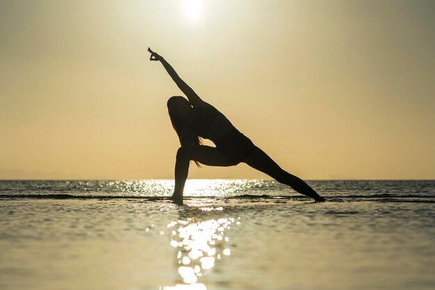 Silhueta de mulher em pose de ioga na praia tropical durante o pôr do sol garota praticando ioga perto da água do mar