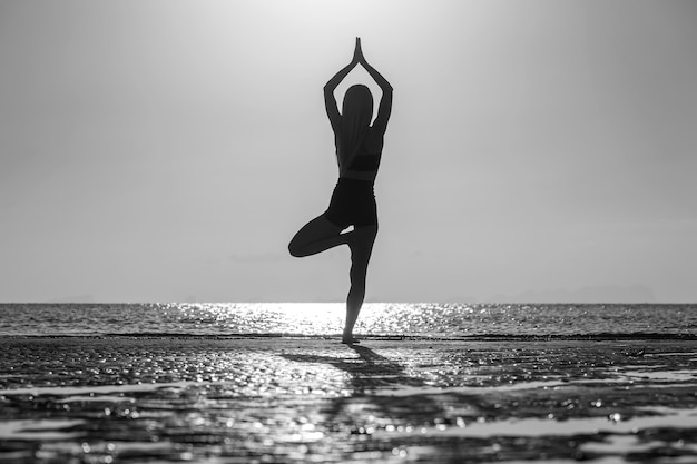Silhueta de mulher em pose de ioga na praia tropical durante o pôr do sol. Menina caucasiana praticando ioga perto da água do mar. Preto e branco